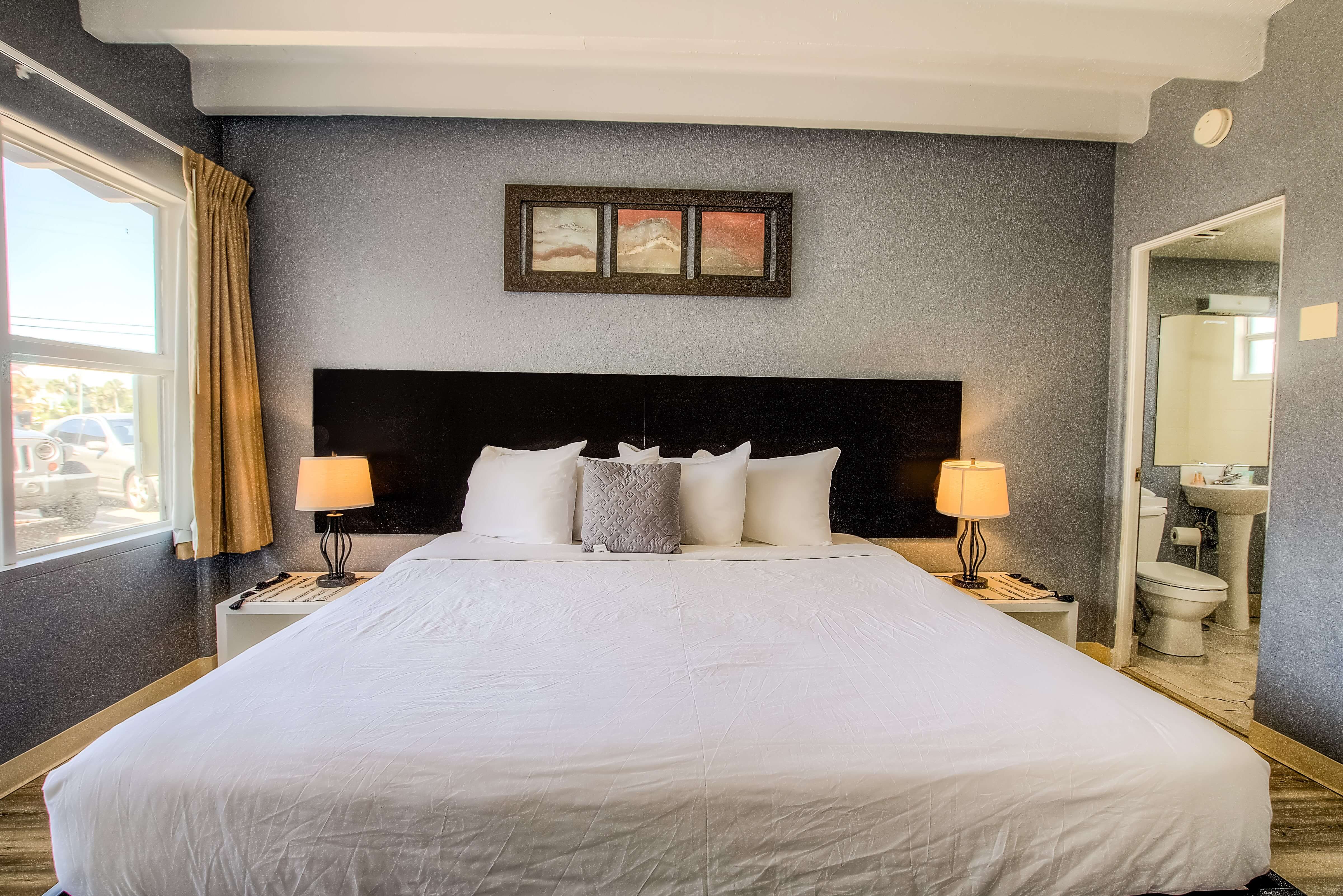 A wide view of a hotel room with a large bed, nightstands, lamps, and a window on the left, with a bathroom doorway on the right.